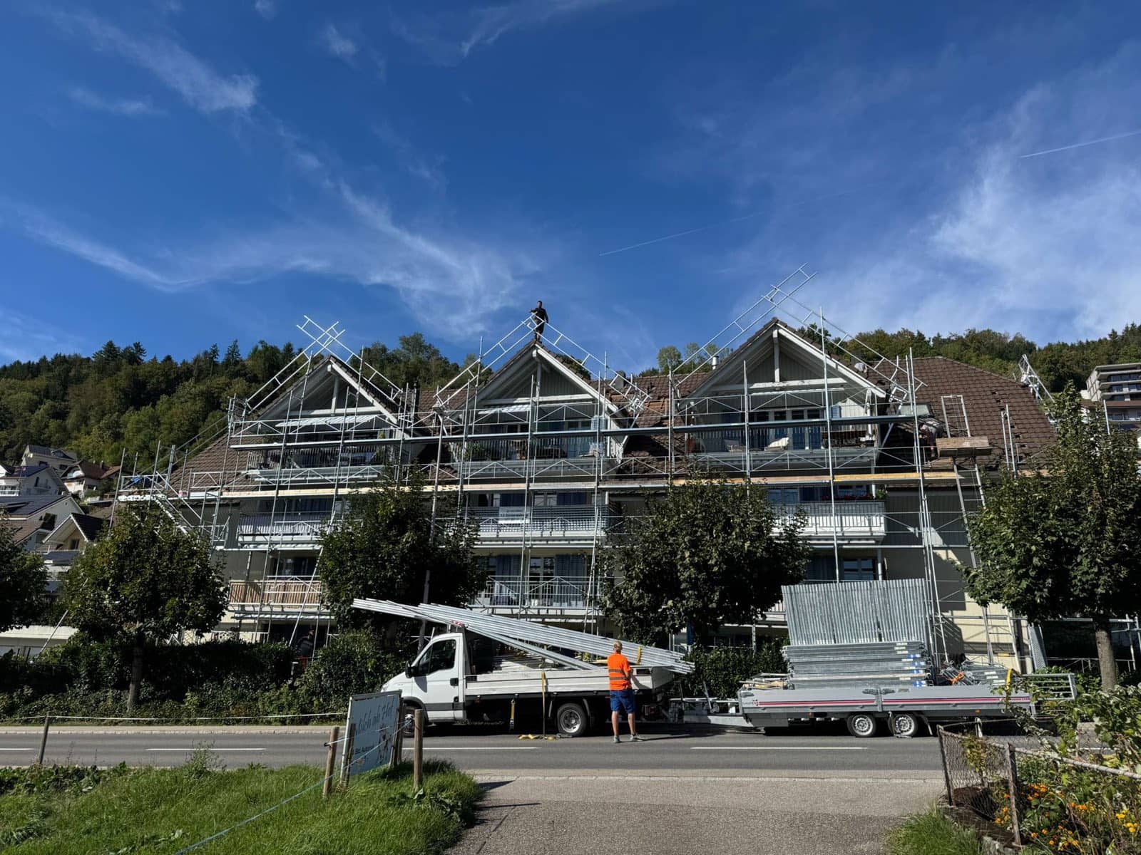 Dachfanggerüst der Elian Gerüstbau GmbH an einem Mehrfamilienhaus mit drei Giebeln während der Dachsanierung, Montagefahrzeug mit Gerüstmaterial im Vordergrund